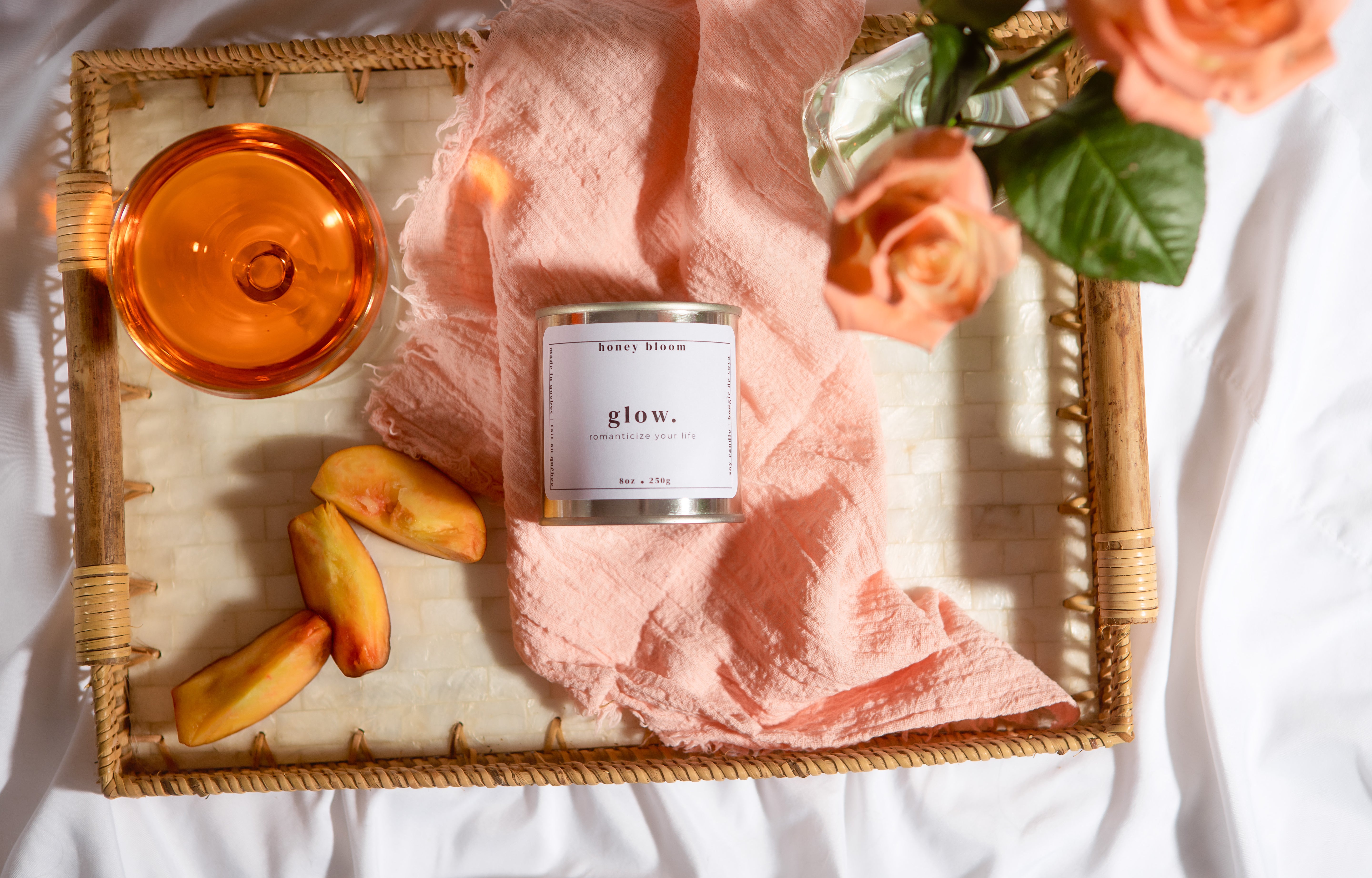 Overhead view of the handmade soy candle “Glow” by Honey Bloom, an artisanal Québec candle brand, styled on a woven tray with peach slices, roses, soft linens, and warm natural light.