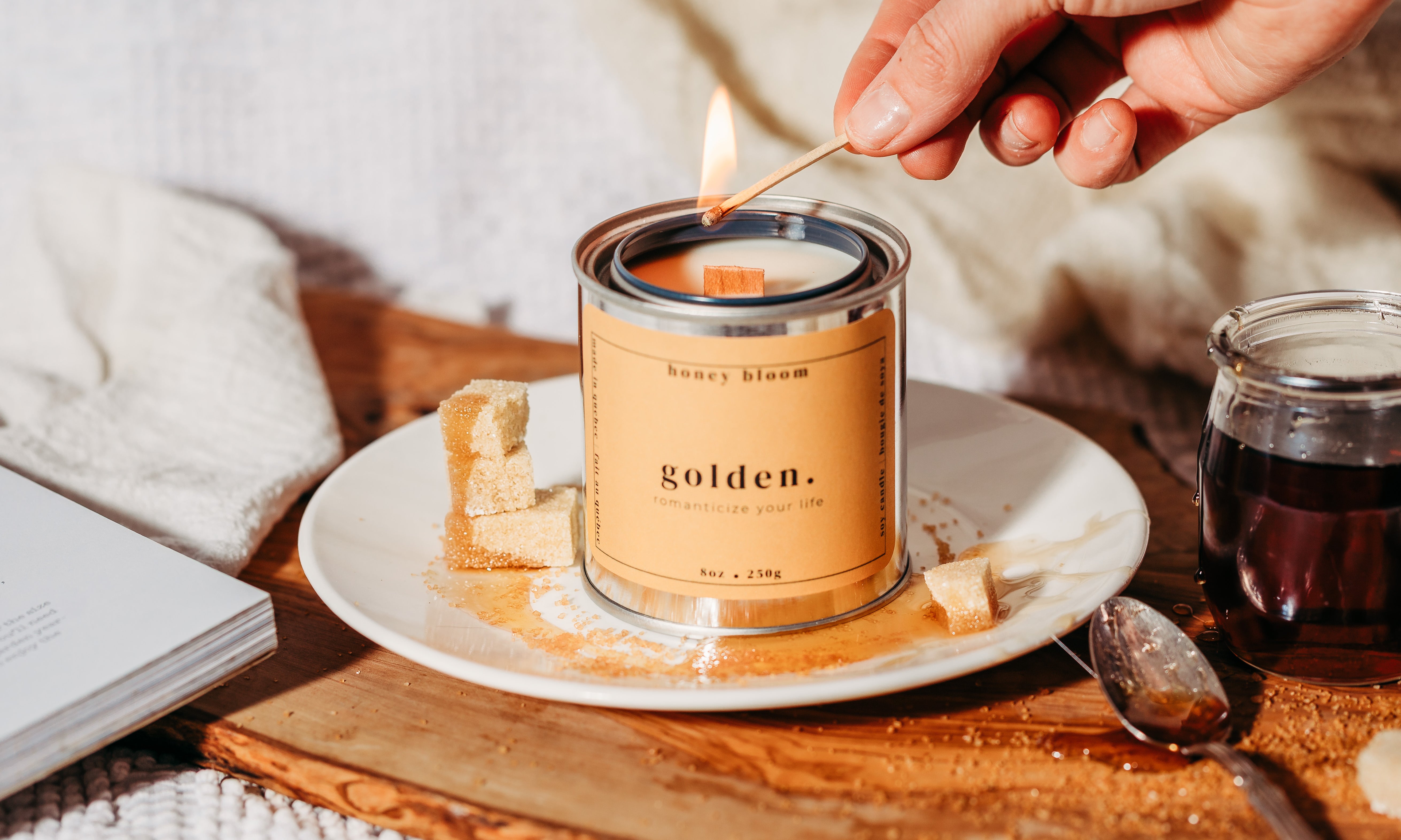 Handmade soy candle “Golden” by Honey Bloom, an artisanal Québec candle brand, being lit by hand on a ceramic plate with brown sugar cubes, tea, and warm natural light.