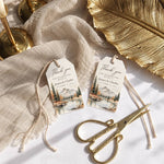 Two personalized wedding thank-you tags with a mountain lake illustration, styled on a white surface with beige fabric, gold scissors, and gold candlesticks.