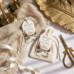 Two personalized thank-you gift tags with a mountain lake illustration are displayed on a beige textured fabric pouch and ribbon, styled with gold scissors, brass candlesticks, and a gold leaf on a white surface.