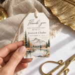 A hand holds a personalized thank you gift tag featuring a scenic mountain lake illustration. The white paper tag has elegant black script text and is styled on a white surface with neutral fabric, gold scissors, and a gold decorative leaf.