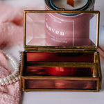Bronze jewelry box with a glass lid on a surface with a pearl necklace and candle in the background.