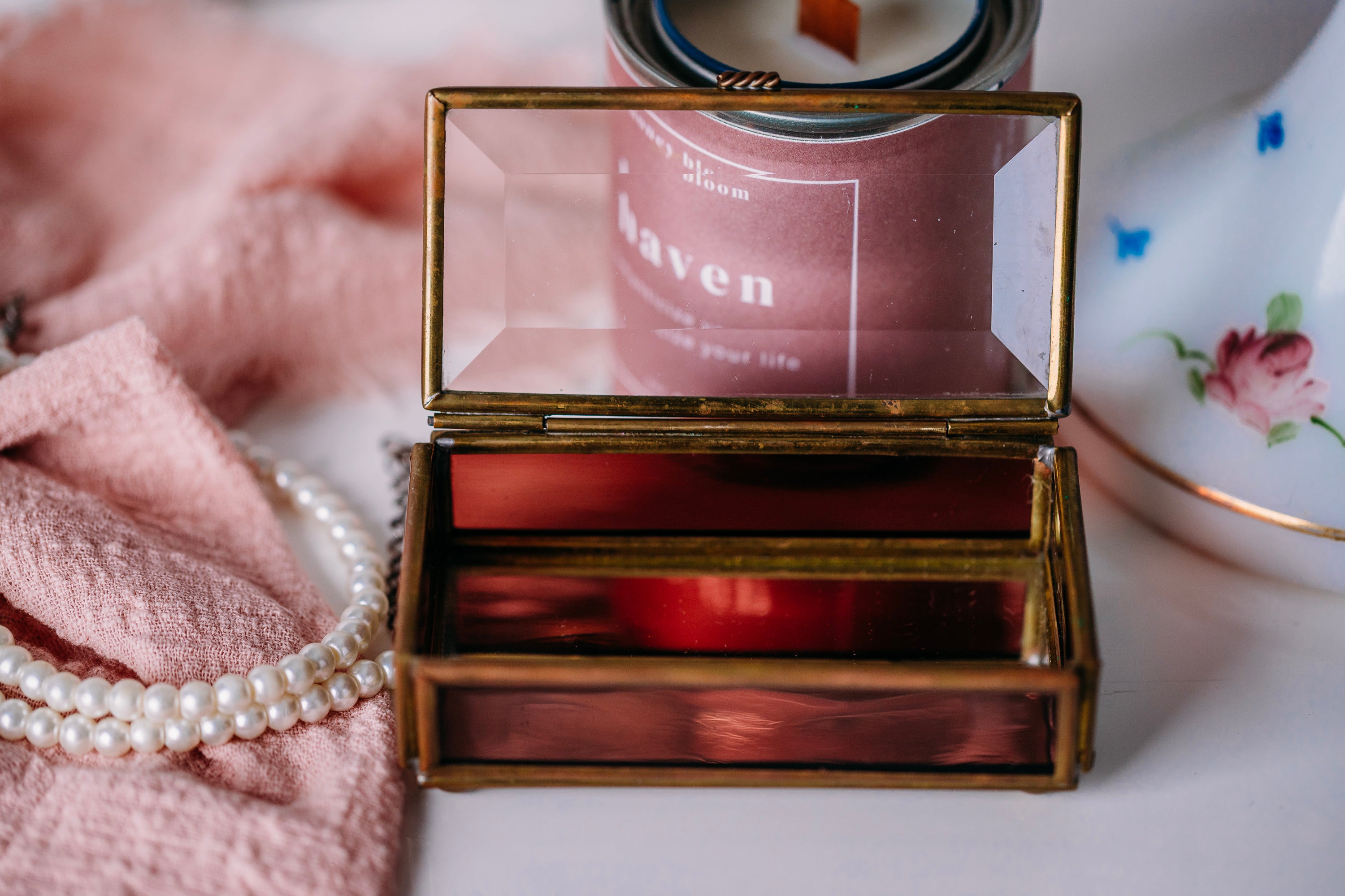 Bronze jewelry box with a glass lid on a surface with a pearl necklace and candle in the background.