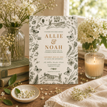 A botanical-themed wedding invitation card is displayed on a rustic wooden table surrounded by decorative props, including a lit candle in a glass holder, stacked books, small vases of white flowers, and a cream fabric runner.