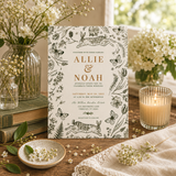 A botanical-themed wedding invitation card is displayed on a rustic wooden table surrounded by decorative props, including a lit candle in a glass holder, stacked books, small vases of white flowers, and a cream fabric runner.