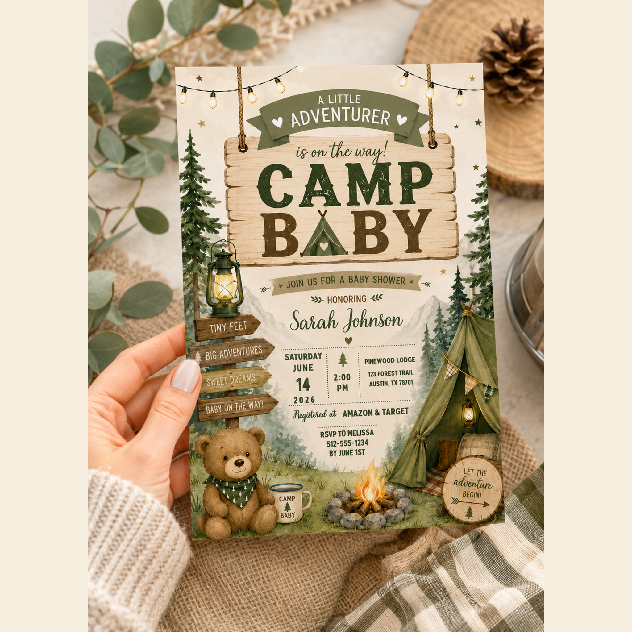 Hand holding a camping-themed baby shower invitation with forest green lettering and illustrated woodland details.