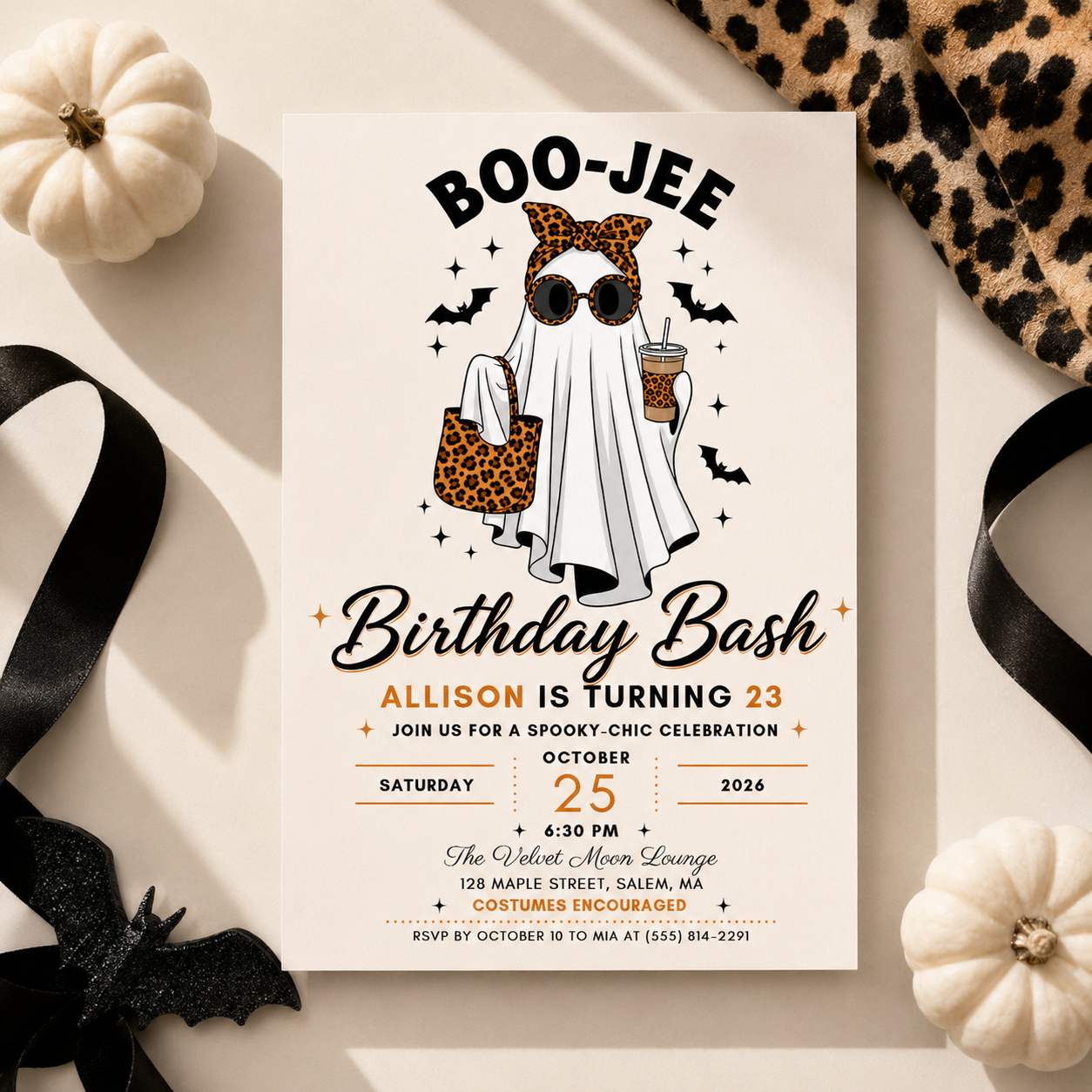 A Halloween-themed birthday invitation card styled in a flat lay with white pumpkins, black ribbons, a leopard-print fabric, and a black bat decoration on a light beige background. The card features a ghost illustration with leopard-print sunglasses and a leopard-print bag, with text reading 'BOO-JEE Birthday Bash' and event details.