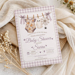 A baby shower invitation card with a lavender and white gingham border, featuring a Halloween-themed illustration of a baby outfit hanging on a clothesline with a ghost, spider, and moon. The card is styled on a neutral surface with beige fabric and dried flowers.