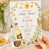 Hand holding a floral honey bee baby shower invitation with personalized coral lettering on a cream background.