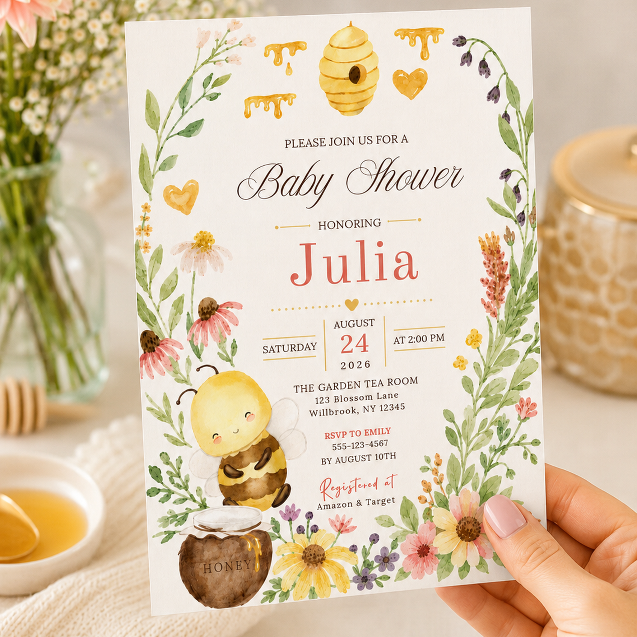 Hand holding a floral honey bee baby shower invitation with personalized coral lettering on a cream background.