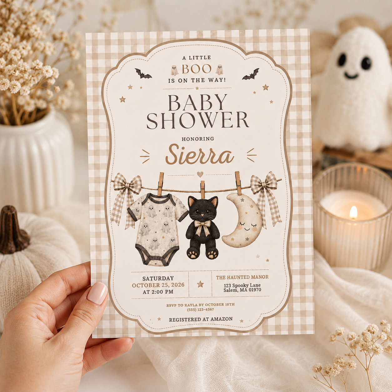 Hand holding a neutral Halloween baby shower invitation with a scalloped frame, gingham bows and a miniature clothesline illustration.