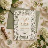 A styled flat-lay of a wedding invitation card with botanical and animal illustrations, placed on top of an olive green envelope. The scene is decorated with white flowers, green leaves, a wooden stamp handle, and a wax seal on a soft beige fabric background.