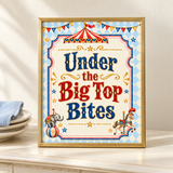 Gold-framed “Under the Big Top Bites” circus food table sign beside ivory plates and a pale blue napkin.