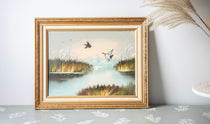 Framed painting of ducks on a lake with reeds on a light gray surface.