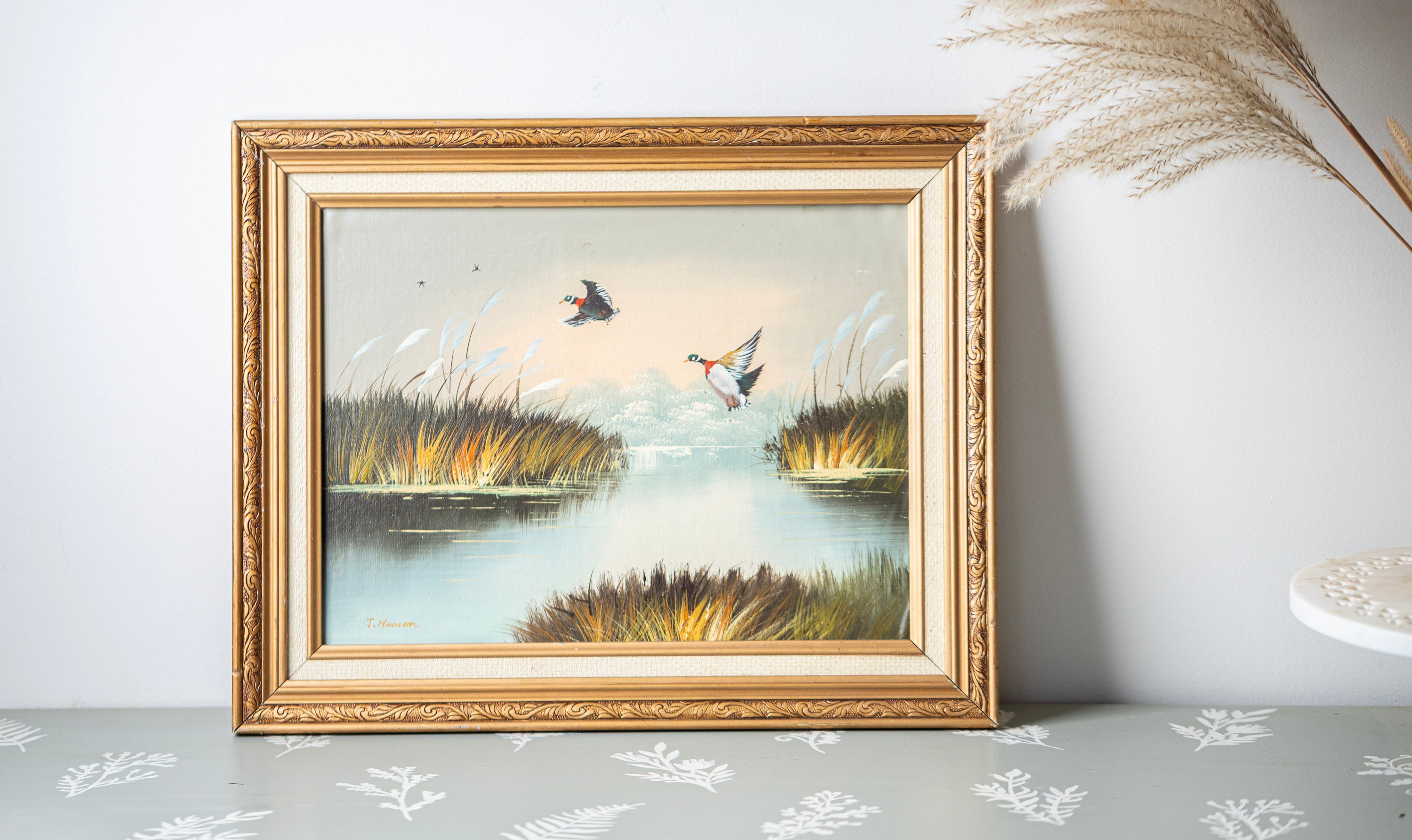 Framed painting of ducks on a lake with reeds on a light gray surface.
