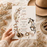 A hand holds a rustic western-themed wedding invitation card featuring a horse, cowboy hat, rope, and dried floral illustrations. The card is styled on a textured cream fabric with a straw hat and dried flowers in the background.