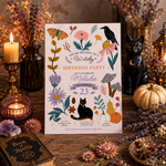 A witch-themed birthday party invitation card is displayed on a rustic wooden table surrounded by seasonal props, including a black cat figurine, dried flowers, candles, and a small black book.