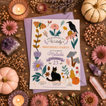 A flat-lay of a witch-themed birthday party invitation card with a lavender envelope, surrounded by autumn and witch motifs including a black cat, mushrooms, a raven, pumpkins, dried flowers, crystals, and candles on a wooden surface.