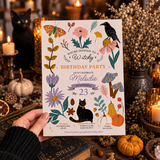 A hand holds a fall-themed 23rd birthday invitation card featuring illustrations of a black cat, crows, pumpkins, flowers, and leaves. The background is a cozy, styled autumn scene with lit candles, books, dried flowers, and pumpkins.