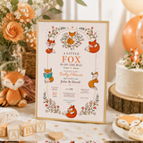 Fox baby shower invitation in a gold frame, displayed beside a cake, peach roses and a knitted fox toy.