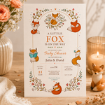 Woodland fox baby shower invitation standing on a rustic table beside peach flowers, a candle and a fox figurine.