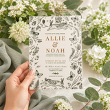 A hand holds a wedding invitation card featuring black botanical illustrations of a deer, butterflies, and foliage on a cream background. The card has elegant gold text reading 'ALLIE & NOAH' and wedding details.
