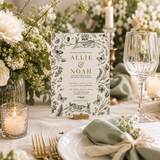 A wedding invitation card displayed on a small wooden stand on a decorated table setting. The card features black botanical illustrations of flowers, leaves, and butterflies on a white background, with the names 'ALLIE & NOAH' in elegant gold lettering.