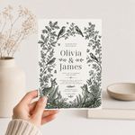 Hand holding a woodland wedding invitation featuring birds, a deer, rabbit and fox in a forest-green floral border.