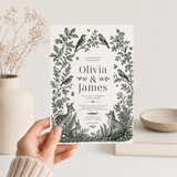 Hand holding a woodland wedding invitation featuring birds, a deer, rabbit and fox in a forest-green floral border.
