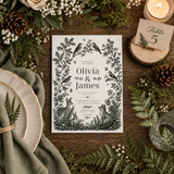 A styled flat-lay of a forest-themed wedding invitation card featuring black botanical and animal illustrations on a white background, surrounded by green ferns, pinecones, a candle, plates, and a table number card on a dark wood surface.